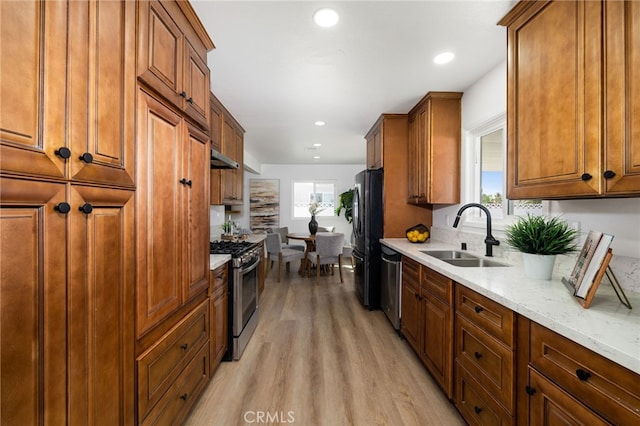 kitchen featuring light stone counters, stainless steel appliances, light wood-style floors, brown cabinetry, and a sink