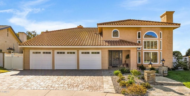 mediterranean / spanish-style house featuring an attached garage, a tiled roof, decorative driveway, stucco siding, and a chimney