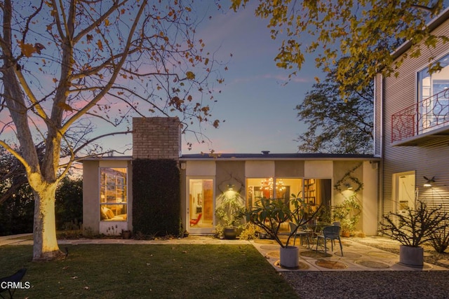 back of property at dusk featuring a patio area, a lawn, a chimney, and stucco siding