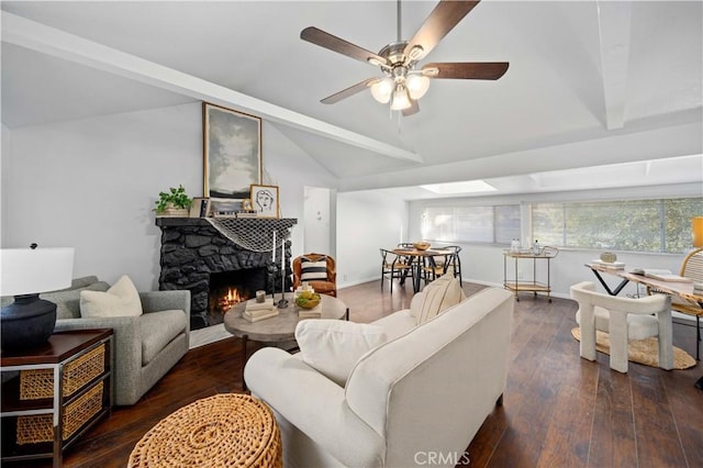 living room with vaulted ceiling with beams, ceiling fan, a stone fireplace, dark wood-style flooring, and baseboards