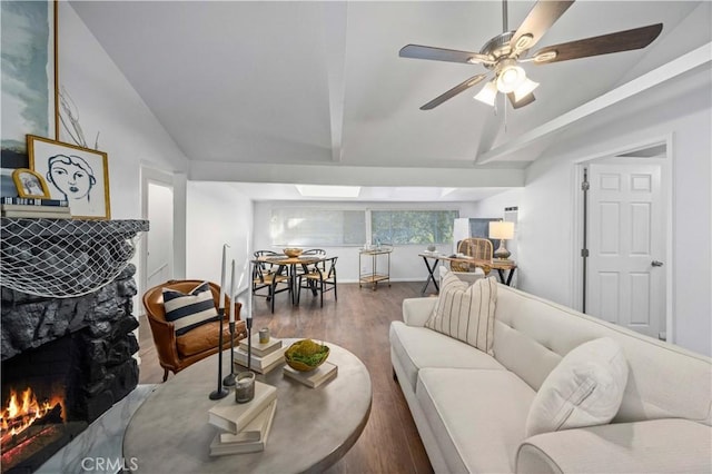 living area featuring lofted ceiling with beams, ceiling fan, wood finished floors, a warm lit fireplace, and baseboards