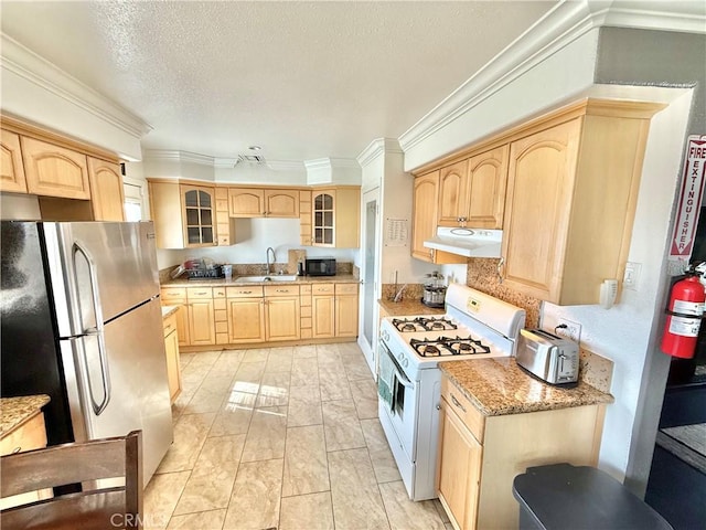 kitchen with light brown cabinets, under cabinet range hood, white range with gas stovetop, freestanding refrigerator, and glass insert cabinets