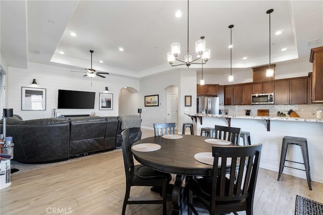 dining area featuring a ceiling fan, arched walkways, a raised ceiling, and light wood finished floors