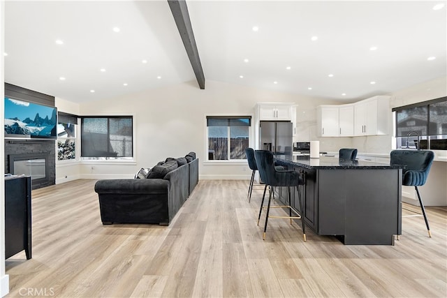 living room featuring vaulted ceiling with beams, light wood-type flooring, a glass covered fireplace, and baseboards