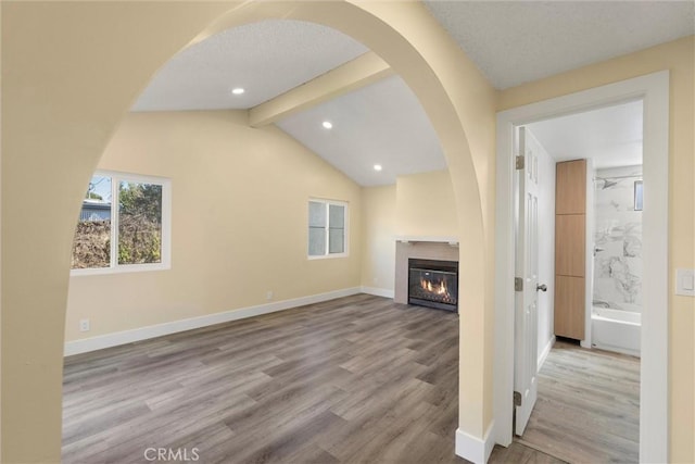 unfurnished living room with wood finished floors, baseboards, vaulted ceiling with beams, arched walkways, and a glass covered fireplace