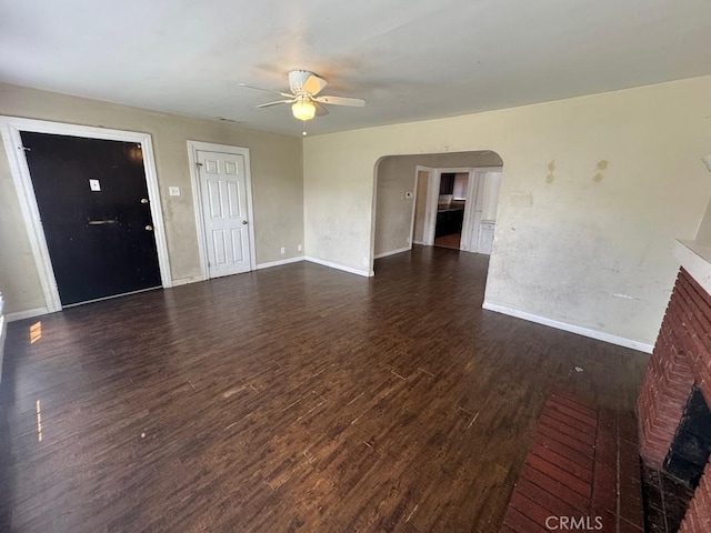 unfurnished living room featuring arched walkways, dark wood-type flooring, a fireplace, a ceiling fan, and baseboards