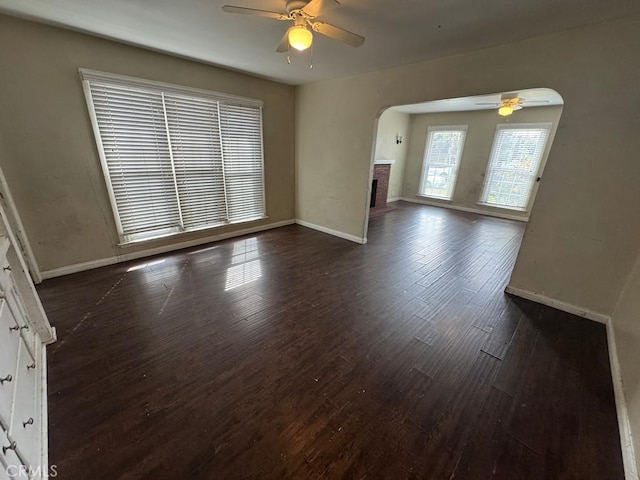 unfurnished living room with baseboards, arched walkways, dark wood-style floors, ceiling fan, and a fireplace