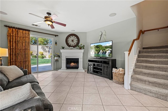 living area featuring light tile patterned floors, ceiling fan, visible vents, stairs, and a glass covered fireplace