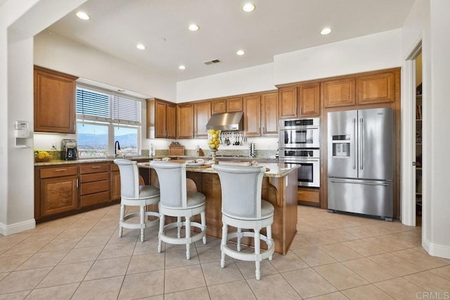 kitchen featuring a center island, appliances with stainless steel finishes, brown cabinetry, light stone countertops, and under cabinet range hood