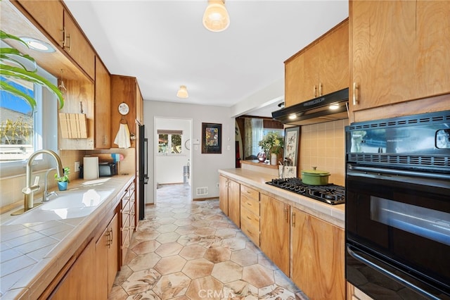 kitchen with tile countertops, under cabinet range hood, a sink, black appliances, and tasteful backsplash