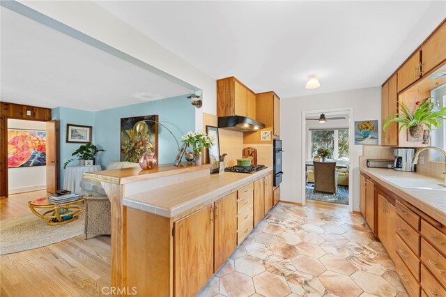 kitchen with a peninsula, a sink, backsplash, tile counters, and under cabinet range hood