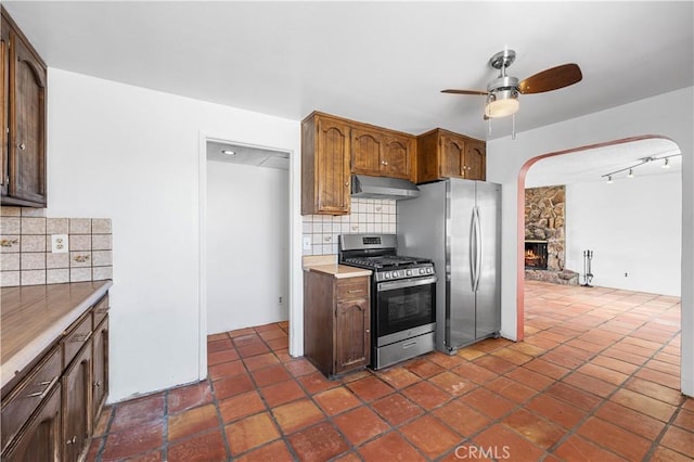 kitchen featuring ceiling fan, stainless steel appliances, under cabinet range hood, a fireplace, and backsplash