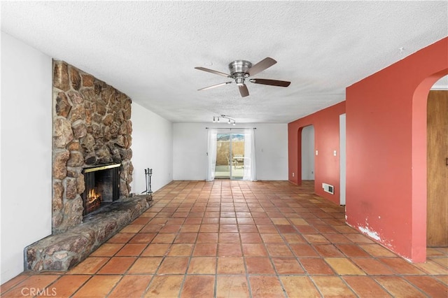 unfurnished living room with arched walkways, visible vents, ceiling fan, a stone fireplace, and a textured ceiling