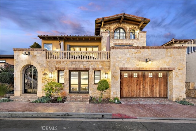 mediterranean / spanish-style home featuring an attached garage, a balcony, stone siding, a tiled roof, and decorative driveway