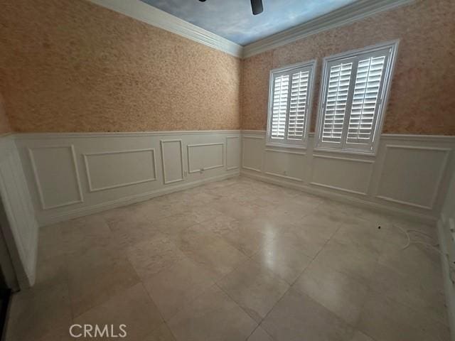 empty room featuring wallpapered walls, ceiling fan, crown molding, and a wainscoted wall