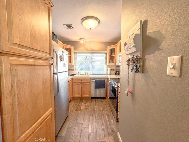 kitchen with stainless steel appliances, light countertops, light brown cabinetry, glass insert cabinets, and a sink