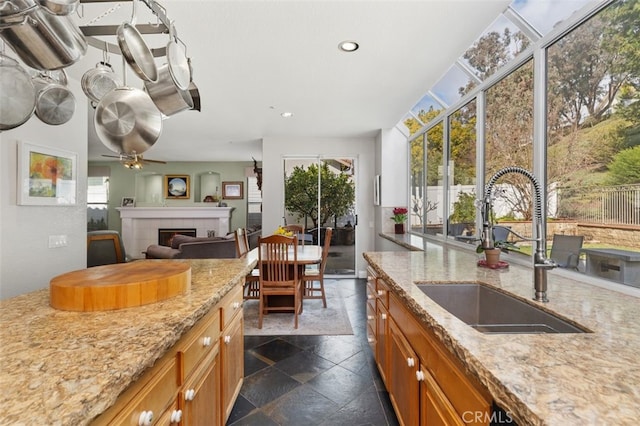 kitchen featuring stone tile floors, a tiled fireplace, brown cabinetry, a sink, and light stone countertops