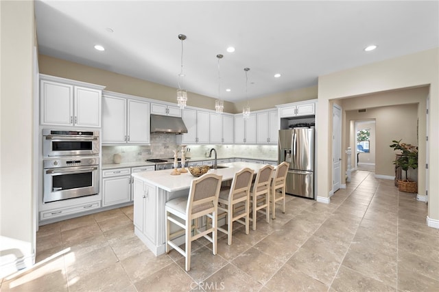 kitchen featuring appliances with stainless steel finishes, decorative light fixtures, a kitchen island with sink, light countertops, and under cabinet range hood