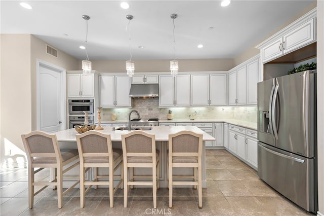 kitchen with under cabinet range hood, stainless steel appliances, light countertops, an island with sink, and decorative light fixtures