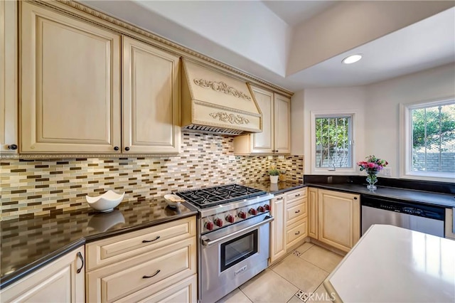 kitchen with appliances with stainless steel finishes, cream cabinetry, custom exhaust hood, decorative backsplash, and dark countertops