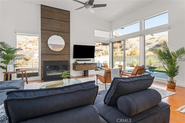 living room featuring ceiling fan, a high ceiling, wood finished floors, baseboards, and a glass covered fireplace