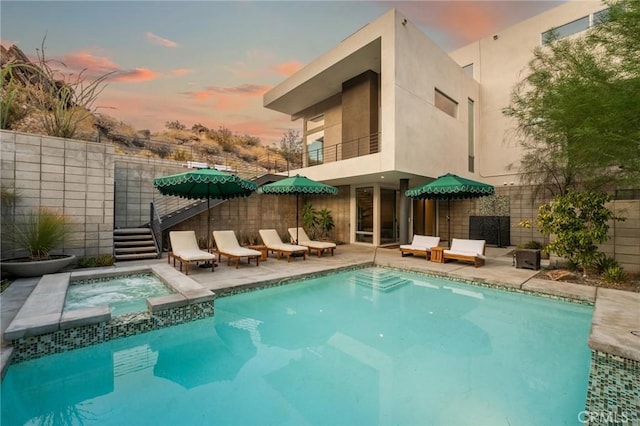 pool at dusk featuring a pool with connected hot tub, fence, and a patio