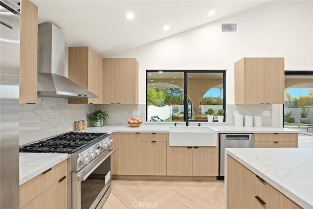 kitchen featuring appliances with stainless steel finishes, vaulted ceiling, wall chimney range hood, light brown cabinets, and a sink