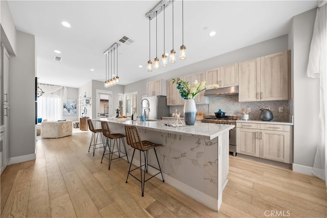 kitchen with decorative backsplash, appliances with stainless steel finishes, light brown cabinets, a large island with sink, and under cabinet range hood