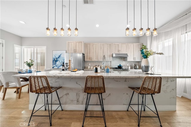 kitchen featuring backsplash, stainless steel fridge, a large island with sink, under cabinet range hood, and a kitchen bar