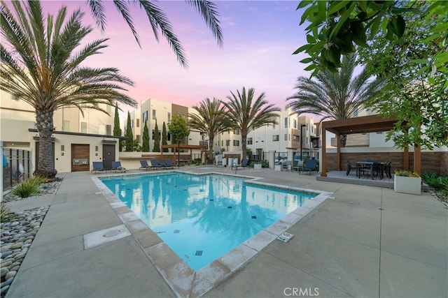 pool at dusk with a patio area, fence, and a community pool