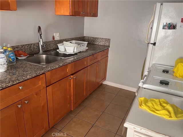 kitchen with brown cabinets, dark stone countertops, freestanding refrigerator, a sink, and light tile patterned flooring