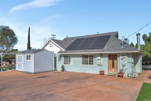 rear view of property with a storage shed, roof with shingles, an outdoor structure, a patio area, and roof mounted solar panels