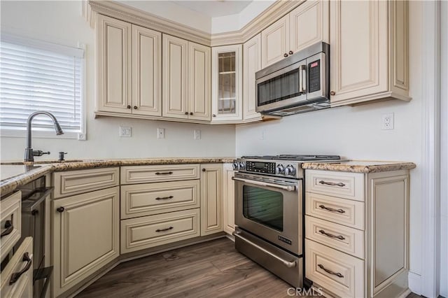 kitchen with cream cabinets, appliances with stainless steel finishes, glass insert cabinets, a sink, and light stone countertops