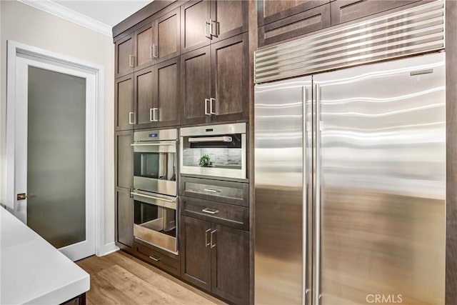 kitchen featuring dark brown cabinetry, light wood finished floors, appliances with stainless steel finishes, ornamental molding, and light countertops