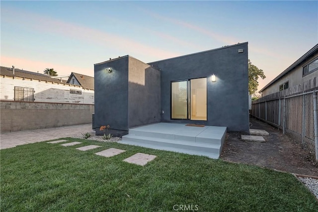 back of house at dusk with stucco siding, a patio, a yard, and fence
