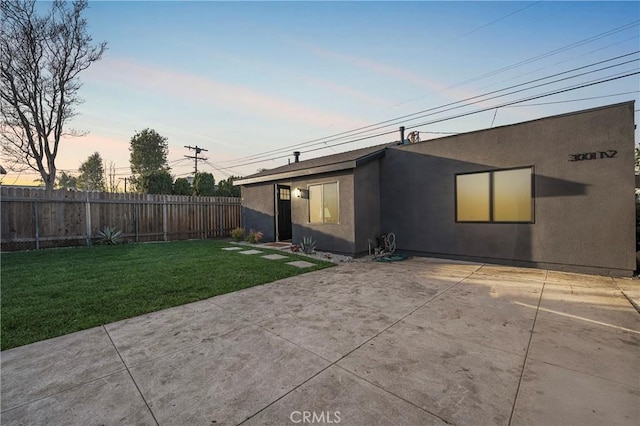 back of house at dusk with a patio area, a yard, fence, and stucco siding
