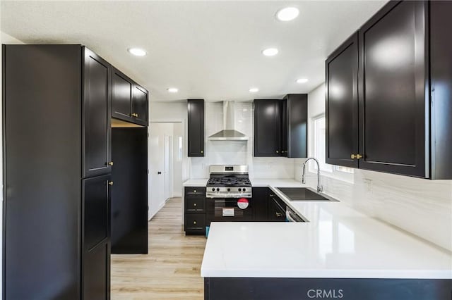 kitchen with light wood finished floors, a sink, stainless steel range with gas cooktop, wall chimney exhaust hood, and light countertops