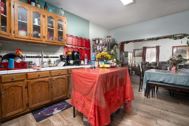 kitchen featuring light wood-type flooring, tile counters, brown cabinets, and glass insert cabinets