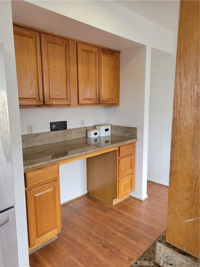 kitchen with brown cabinetry, wood finished floors, built in desk, and freestanding refrigerator