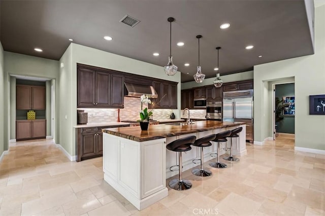 kitchen featuring wall chimney range hood, a sink, appliances with stainless steel finishes, visible vents, and dark brown cabinetry