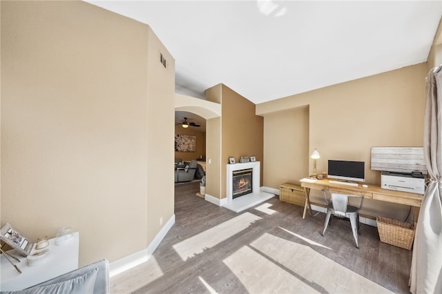 office space with arched walkways, wood finished floors, a ceiling fan, baseboards, and a glass covered fireplace