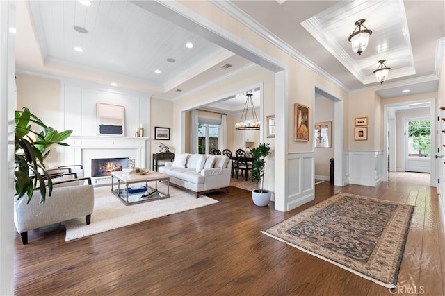 living room featuring a warm lit fireplace, wood-type flooring, a tray ceiling, a decorative wall, and a notable chandelier