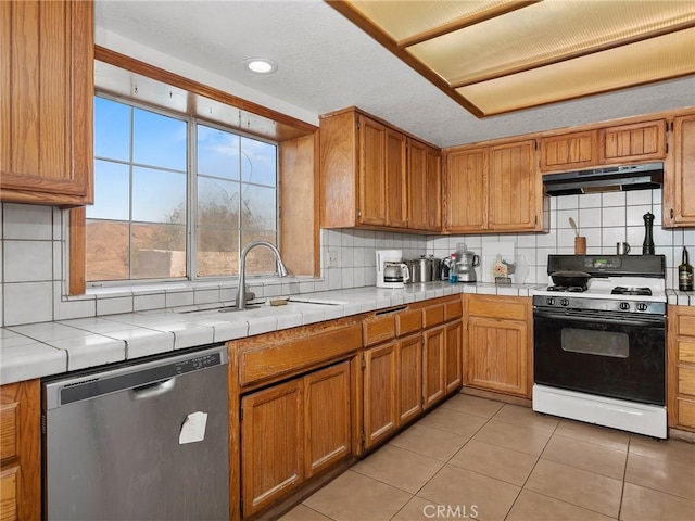 kitchen with tile countertops, under cabinet range hood, a sink, stainless steel dishwasher, and gas stove