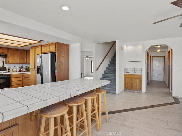 kitchen with ceiling fan, white gas range oven, tile counters, stainless steel fridge, and a kitchen bar