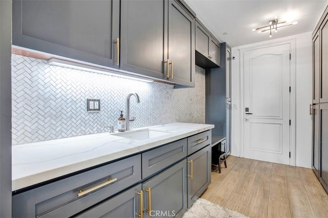 kitchen featuring gray cabinetry, a sink, backsplash, light stone countertops, and light wood finished floors