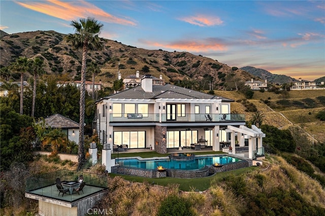 back of property at dusk with a chimney, a pool with connected hot tub, a patio area, a mountain view, and a balcony