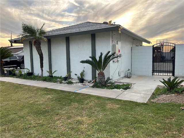 property exterior at dusk with a shingled roof, fence, a lawn, a gate, and stucco siding