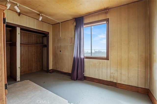 unfurnished bedroom featuring wood walls, baseboards, finished concrete flooring, a closet, and rail lighting