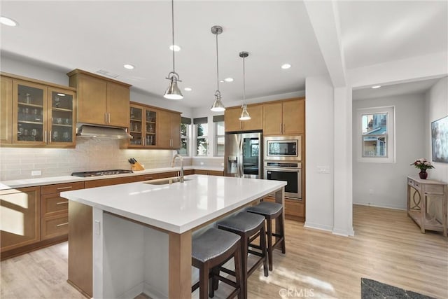 kitchen featuring an island with sink, appliances with stainless steel finishes, light countertops, under cabinet range hood, and a sink