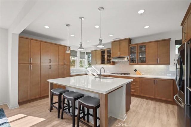 kitchen with a kitchen island with sink, stainless steel appliances, light countertops, brown cabinetry, and glass insert cabinets